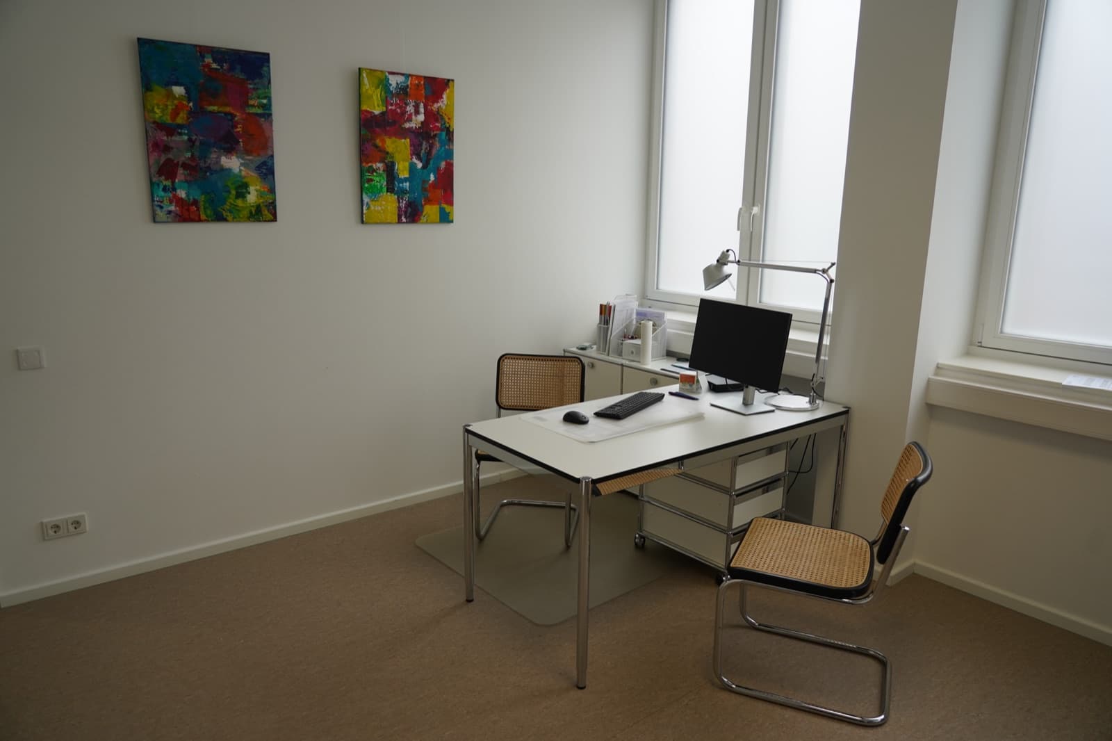 Sprechzimmer mit Schreibtisch, Patientenstuhl und bunter Kunst